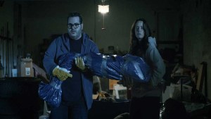 Kaya Scodelario and Josh Gad standing in a garage holding a body wrapped in plastic in a still from Alex Winter's "Aduthood."