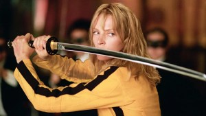 A woman with blonde hair wearing a yellow and black jacket is holding a samurai sword with both hands, aimed forward. The background is blurred, suggesting an indoor setting with other figures present. The individual appears focused and determined.